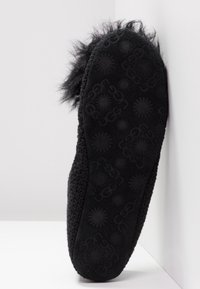 Chausson noir avec semelle texturée présentant des logos UGG et des motifs soleil, dessus en maille tricotée, et bordure en fourrure moelleuse au talon.