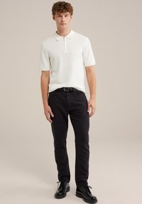 Rövid ujjú, galléros fehér kötött polóing fekete farmernadrággal és fekete fűzős cipővel kombinálva, cipzáras nyakkal és texturált anyaggal.