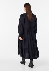 Largo vestido negro en capas con mangas largas y un escote redondo. Hecho de una tela ligera, con una silueta fluida y un brillo sutil.