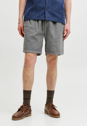 Homme portant un short en velours côtelé gris, des chaussettes marron foncé et des chaussures bateaux en cuir marron, debout devant un fond clair uni.