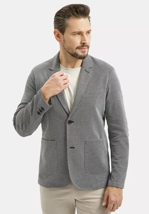 Mężczyzna poprawiający kołnierz szarej, fakturowanej marynarki na białej koszuli, ubrany w beżowe spodnie, patrzący w prawo na jednolitym tle.