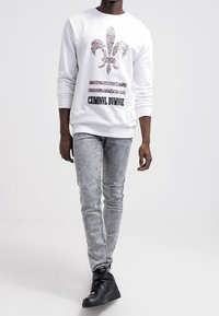 Bílá mikina s květinovým motivem fleur-de-lis, černým textem a pruhy. Kombinováno se světlě šedými džíny a černými teniskami s vysokým svrškem.