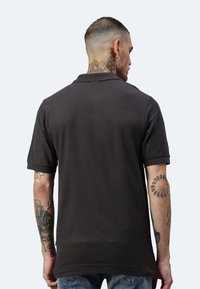 Dunkelgraues Poloshirt mit Kragen und kurzen Ärmeln, aus einem strukturierten Stoff. Auffällige Tattoos sind an den Armen sichtbar.