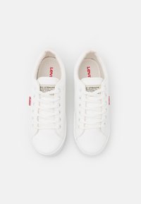 Fehér vászon sneaker, kerek orral, hat fémlyukkal, lapos fehér fűzőkkel, és egy piros Levi's logóval az oldalon.