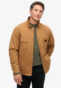Chaqueta de algodón marrón con cuello alzado, botones a presión, dos bolsillos delanteros y un parche de logo, utilizada sobre un suéter verde texturizado.