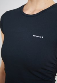Tamsiai mėlyna prigludusi medvilninė marškinėliai, su trumpomis rankovėmis ir apvalia iškirpte, ant krūtinės esantis baltas "Converse" logotipas.
