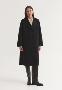 Femme aux cheveux bruns et raides porte un long manteau noir à double boutonnage sur un haut blanc et des bottes noires montantes jusqu'aux genoux, debout devant un fond uni.
