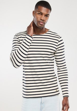 Homme aux cheveux courts portant une chemise à manches longues rayée horizontale blanche et noire, jean bleu clair, touchant son cou.