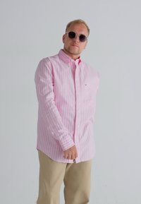 Light pink striped button-up shirt with long sleeves, worn with beige pants. A person wears sunglasses and stands against a plain background.