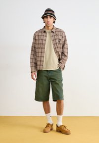 Brown and cream plaid shirt over a beige t-shirt, green denim shorts, white socks, brown shoes, and a black and beige bucket hat.