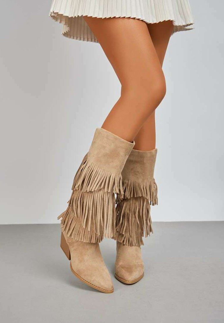 Bottes de cheville en suede beige avec des détails frangés, bout pointu et talon carré. Portées avec une jupe plissée, mettant en valeur la texture des bottes.