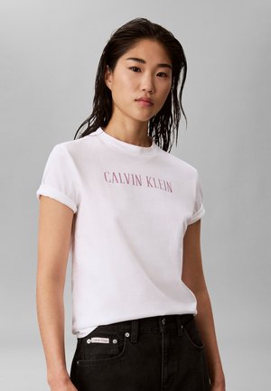 Jeune femme aux cheveux mouillés portant un t-shirt blanc Calvin Klein et un jean noir sur un fond gris uni.