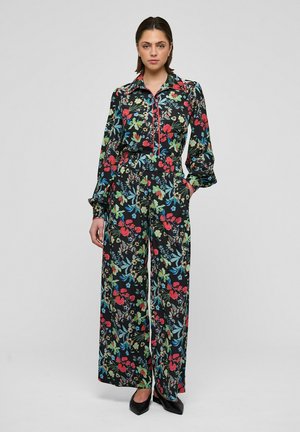 Nainen seisoo yllään musta kukallinen paita ja leveälahkeiset housut, joissa punaisia, vihreitä ja sinisiä kukkia, yhdistettynä mustiin korkokenkiin.