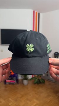 Cappellino da baseball nero con un trifoglio a quattro foglie verde ricamato sulla parte frontale e testo sul lato destro. Materiale in cotone, design strutturato.