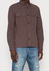 Hnědá košile z vlněné směsi s dlouhými rukávy, dvěma náprsními kapsami a černými knoflíky. Nošená přes opotřebované modré džíny.
