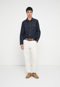 Koszula dżinsowa z kołnierzykiem zapinanym na guziki i dwoma kieszeniami na piersi; w zestawieniu z jasnymi spodniami i brązowymi butami. Gładki kolor i klasyczny design.