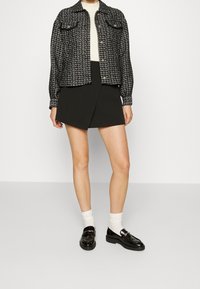 Svart och vit mönstrad jacka med två bröstfickor, kombinerad med en svart omlottkjol och blanka svarta loafers, bärs över vita strumpor.