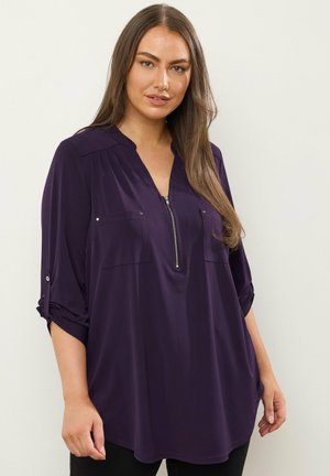 Blouse violette à manches longues avec fermeture éclair sur le devant, deux poches poitrine et manches retroussables. Tissu lisse, coupe décontractée.
