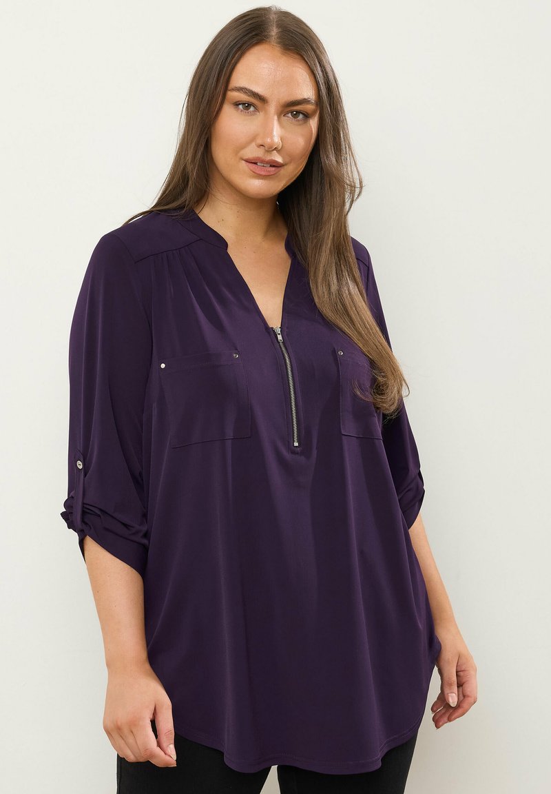 Lila Bluse mit langen Ärmeln und Reißverschluss vorne, zwei Brusttaschen und hochgekrempelten Ärmeln. Glatter Stoff, lässige Passform.