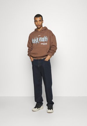 Brun hættetrøje med blå grafisk tekst, mørk jeans og sorte sneakers med cremefarvede detaljer. Uformel pasform, afslappet silhuet og forlommesdetalje.