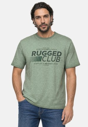 Homme aux cheveux foncés et à la barbe, portant un T-shirt vert avec le texte « "Rugged Club" » et un jean bleu, debout avec une main dans la poche sur fond blanc.