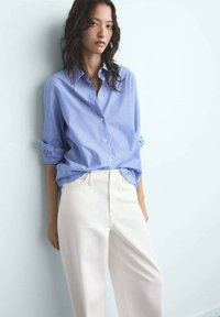 Vrouw in een blauw gestreepte button-up shirt met opgerolde mouwen, gestoken in hoge witte broeken, staande tegen een lichte muur.