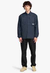 Marineblauwe lichtgewicht jas met drukknopen, elastische manchetten en een voorzak. Gecombineerd met donkere jeans en beige schoenen.