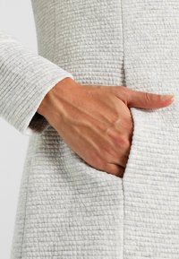 Tejido texturizado de color gris claro con un patrón acolchado, que presenta un bolsillo lateral. Hay una mano colocada dentro del bolsillo, mostrando el material.