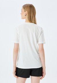 Frau mit blonden Haaren, die ein einfaches weißes T-Shirt mit kurzen Ärmeln und schwarze Shorts trägt, mit dem Rücken zu einer weißen Hintergrund. 