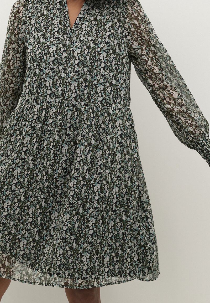 Femme portant une robe noire fleurie à manches longues avec un col en V et une taille froncée.