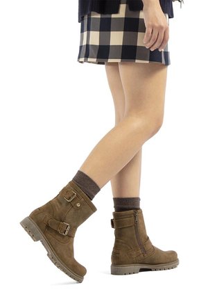 Botas de ante marrón hasta el tobillo con cremallera lateral, detalles de hebilla y suelas texturizadas. Combinadas con calcetines grises y una falda de cuadros.