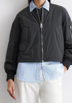 Persoon die een zwarte bomberjack met ritssluiting draagt, over een blauw-wit gestreept overhemd met knopen en witte broek, tegen een neutrale achtergrond.