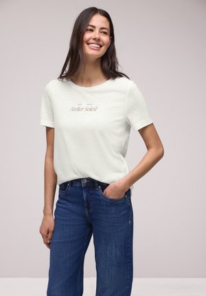 Femme souriante aux cheveux foncés porte un t-shirt blanc à manches courtes « Atelier Soleil » et un jean bleu, debout avec une main dans la poche.