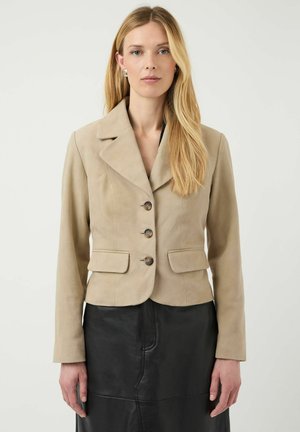 Blonde woman wearing beige buttoned blazer and black skirt, standing against plain light background, looking forward with neutral expression.