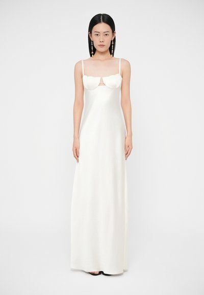 Femme portant une longue robe blanche sans manches avec des bonnets à festons et des bretelles fines, debout devant un fond blanc uni.