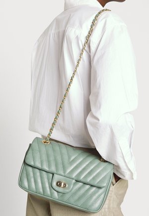 Henkilö, jolla on valkoinen paita ja beiget housut, kantaa mintunvihreää tikattua olkalaukkua, jossa on kultainen ketjuhihna, selässään.