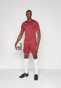 Rode sportshirt met gele logo, bijpassende shorts, zwarte voetbalschoenen en witte sokken. Houdt een zwart-wit voetbalt naar.