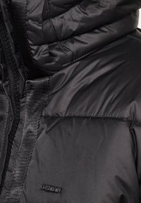 Černá prošívaná bunda s vysokým límcem, hladkou texturou a kontrastním černým zipem s logem na hrudi.