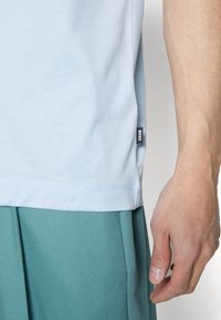 Světle modré bavlněné tričko s bočním štítkem, v kombinaci s texturovanými tyrkysovými šortkami. Částečně viditelná ruka, která ukazuje přirozený tón pleti.