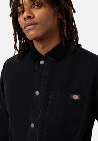 Veste de travail en velours côtelé noir avec fermeture à boutons-pression, poche poitrine avec un patch logo et un col.