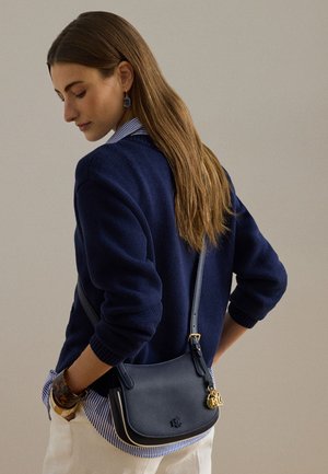 Mujer con cabello largo y castaño, que lleva un suéter azul marino, camisa a rayas, pantalones blancos y un bolso cruzado de cuero azul marino con un charm dorado.