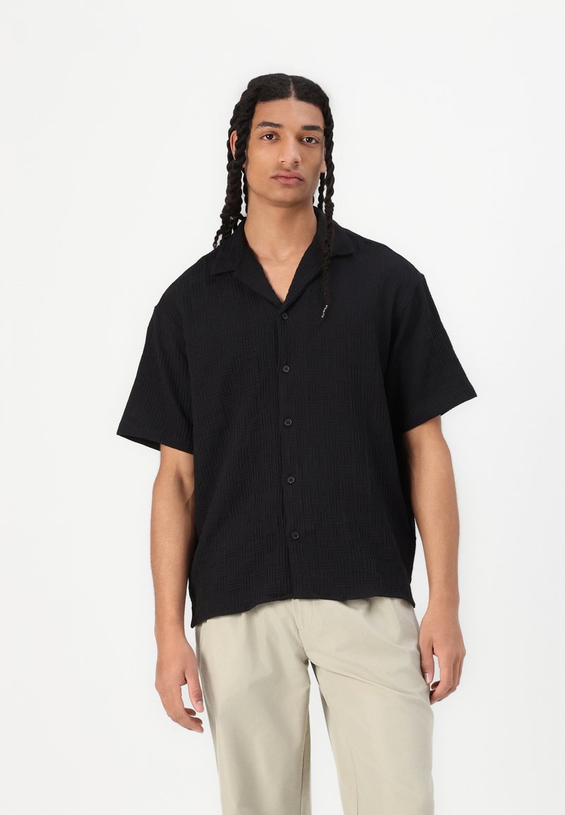 Camicetta nera a maniche corte con un motivo testurizzato e bottoni, con colletto. Indossata con pantaloni beige chiaro.