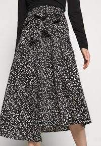 Svart midi kjol med vitt oregelbundet prickmönster och stor rosett knuten i midjan, bärs med en långärmad svart topp.