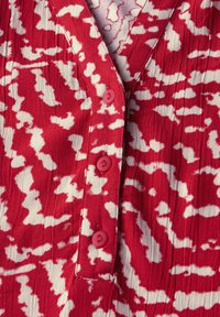 Czerwono-biała tkanina z wzorem, z pionowym przeszyciem, z dekoltem w serek i trzema czerwonymi guzikami na środku przodu.