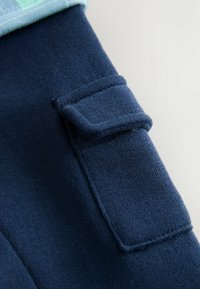 Námornícky modré elastické nohavice z tkaniny s textúrovanou kapsou, mäkkým materiálom a prehnutým svetlomodrým lemom v hornej časti.