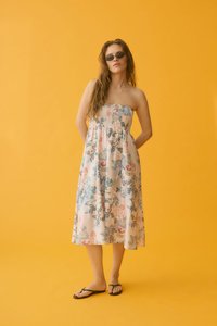 Femme portant des lunettes de soleil foncées, une robe midi fleurie sans bretelles et des sandales noires, debout devant un fond jaune uni.