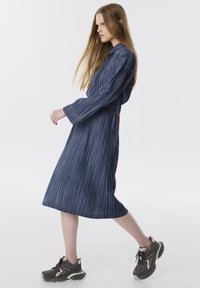 Vestido largo de rayas azules con cintura ajustada y falda fluida. Cuenta con mangas largas y un cuello abotonado. Se lleva con zapatillas oscuras.