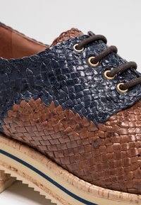 Gros plan d'une chaussure en cuir tressé marron et bleu marine avec des lacets marron foncé et une semelle beige comportant une bande bleue.