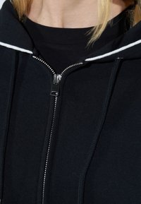 Felpa nera con zip, dotata di una cerniera metallica, con cordini bianchi a contrasto e polsini a costine. Il tessuto appare morbido con una superficie liscia.