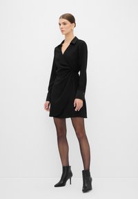 Abito nero avvolgente con maniche lunghe, scollatura a V profonda e dettaglio con fiocco in vita. Abbinato a collant neri trasparenti e stivali alla caviglia.